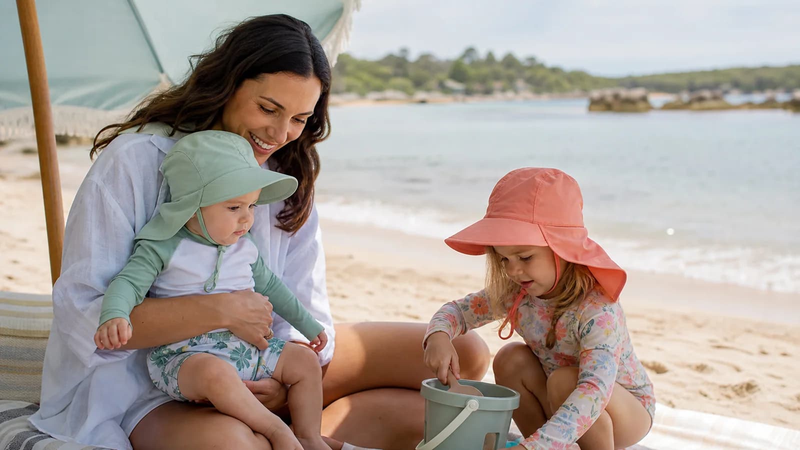 Parent with a baby and toddler wearing full-brim sun hats in shade at the beach